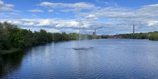 Joki, jonka keskellä suihkulähde. Taustalla teollisuusalue savupiippuineen.