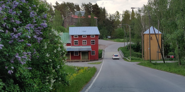 Vasemmalta oikealle: kukassa oleva syreeni, punainen rakennus, asfalttitie, jossa yksi auto ja keltainen rakennus.