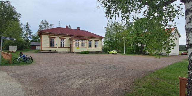 Vasemmalla keltainen puurakennus. Vieressä vihreää puustoa, jonka takana kivinen vaalea rakennus. 
