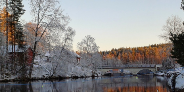 Talvinen maisema, jossa keskellä virtaa joki. Taustalla kaarisilta, jonka toisella puolella punainen puinen rakennus.