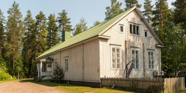 Vaalea hirsitalo, jossa vihreä katto. Taustalla metsää.