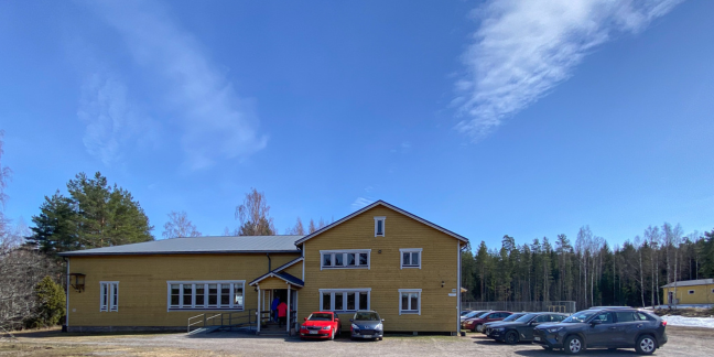 Keltainen puutalo, jonka edessä hiekkakentä, autoja parkissa ja havupuita.