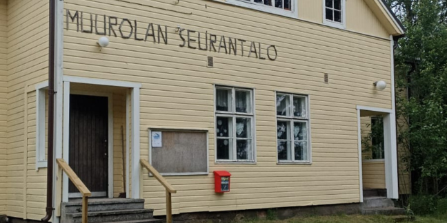 Vaaleankeltainen puinen rakennus, jonka seinässä oksista tehty kyltti "Muurolan seurantalo".