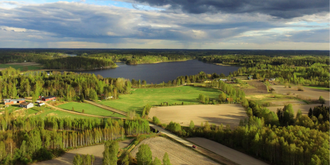 Ilmakuva, jossa näkyy kesäistä maalaismaisemaa, metsiä ja peltoja sekä järvi.