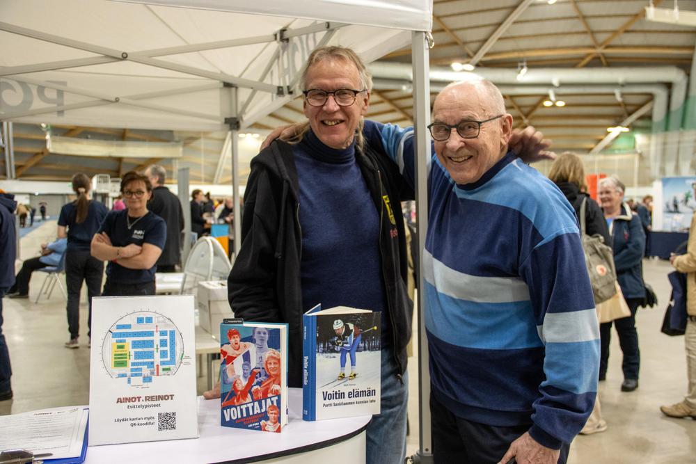 Kaksi henkilöä seisoo tapahtumahallissa esittelypisteellä, pöydällä kirjoja ja esite. Taustalla näkyy muita kävijöitä ja näyttelyrakenteita.