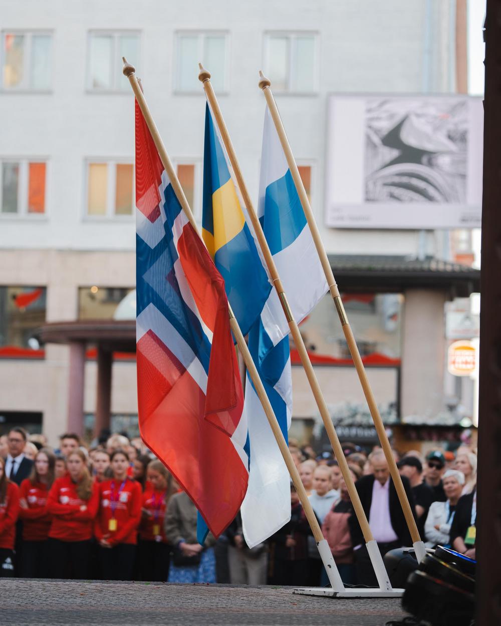Suomen, Ruotsin ja Norjan liput kolmijalassa lipputangossa. Taustalla ihmisiä.