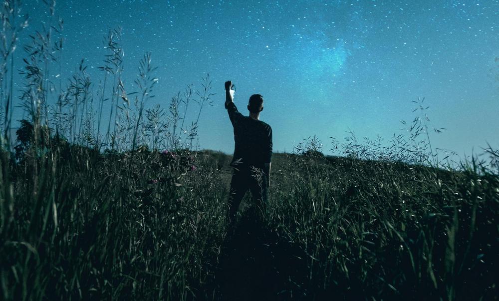 Keskellä kuvaa on nuori mies selin kameraan. Mies katselee öiselle niitylle lyhty kädessään. Yllä kaartuu kirkas tähtitaivas.