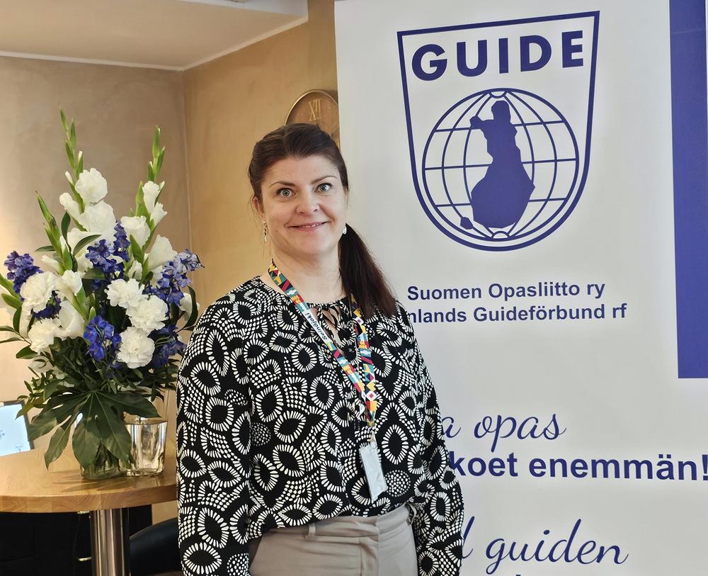 Kuvassa Suomen Opasliiton puheenjohtaja vierellään kukkaasetelma ja taustalla Opasliiton ständi, jossa teksti Ota Opas- koet enemmän