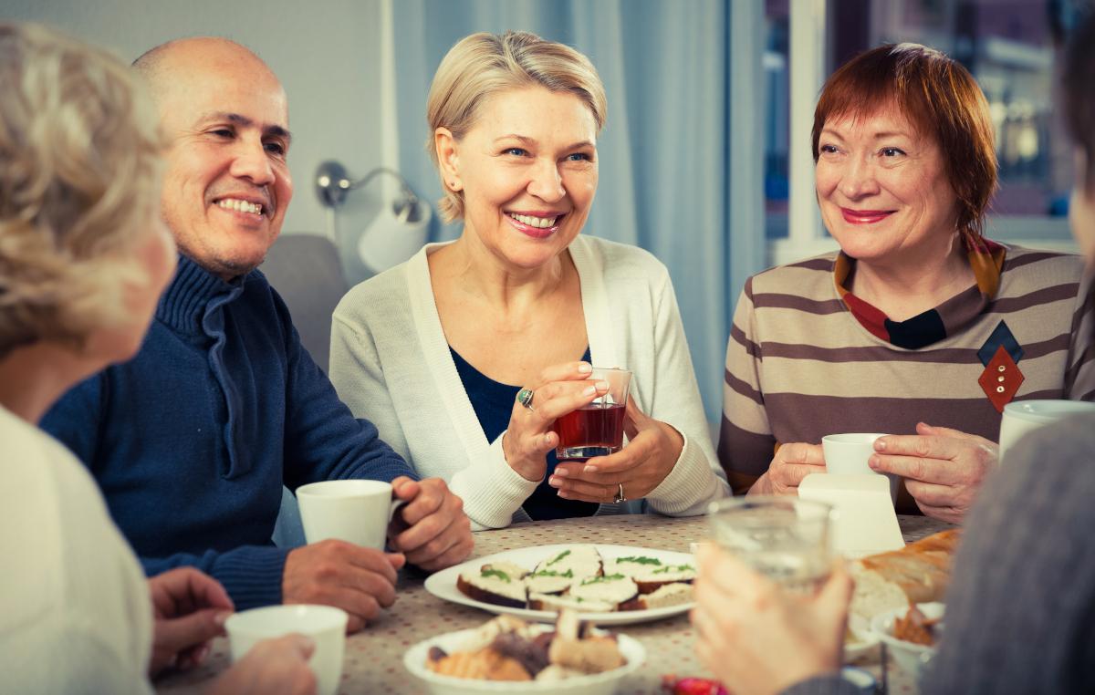 Kuvassa viisi henkilöä istuu pöydän ääressä kahvilla/teellä. Kaksi naista ja yksi mies hymyilee kasvot kameraan päin. Pöydällä on voileipiä lautasella, keksejä kipossa ja pullapitko.