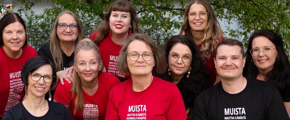 Koko Muistiyhdistyksen iloinen henkilökunta vihreässä ympäristössä Muistiyhdistyksen toimiston pihalla, mustissa ja punaisissa, samanlaisissa t-paidoissa