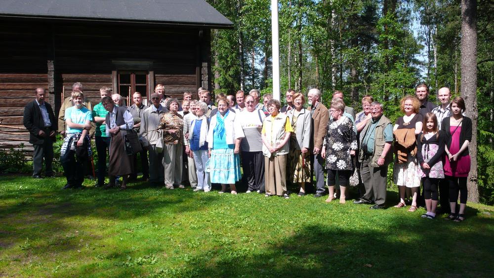37 henkilöä puutalon pihapiirissä vihreällä nurmella, metsää taustalla.
