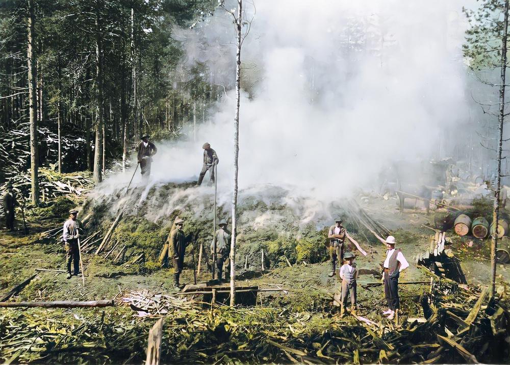 Savuava kumpu, jonka ympärillä ja päällä 1900-luvun alun työmiesten tyyliin pukeutuneita miehiä. Muutama tervatynnyri ympärillä. Väritetty mustavalkokuva.