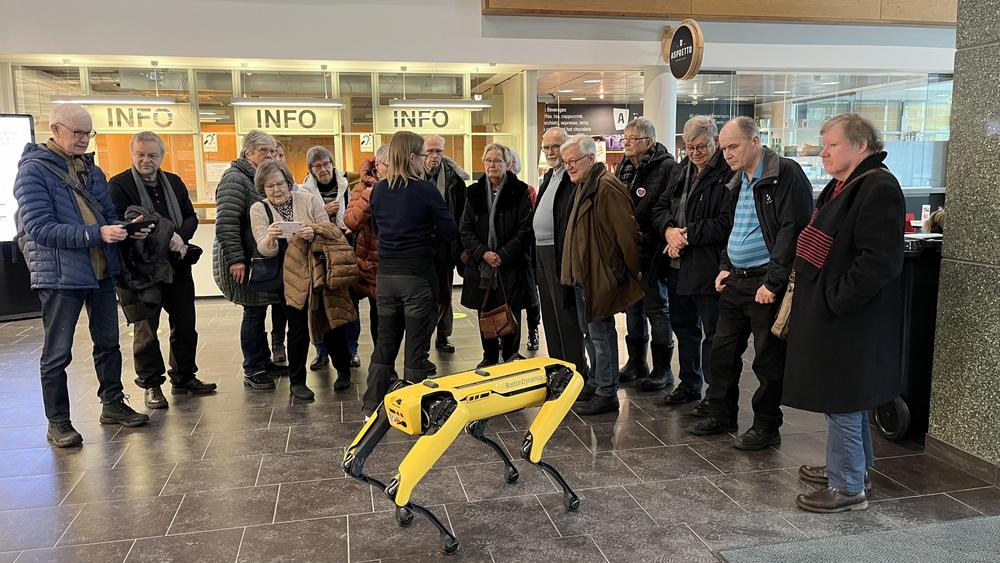 15 vertaisohjaajaa seisoo yliopiston aulassa katsomassa keltaista robottikoiraa.