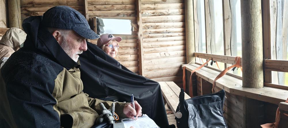 Mies ja nainen Liminganlahden lintutornin ensimmäisessä kerroksessa katselemassa merelle. Molemmilla päällään sadevaatteet.