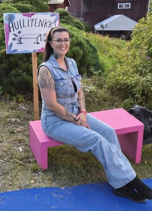 Kuvassa on blogin kirjoittaja Heli Toivonen, joka istuu matalalla pinkillä penkillä. Helin pään takana näkyy käsin maalattu kyltti, jossa lukee Huilipenkki. Helillä on silmälasit, tummanruskeat pitkät hiukset ja valkoinen hiuspanta. Hän katsoo kuvaajaa kohti ja hymyilee.