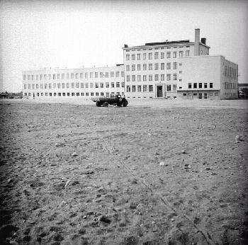 Mustavalkoinen kuva, jossa hiekkakenttä ja sen takana näkyvä koulurakennus, sen edessä kuorma-auto. 
