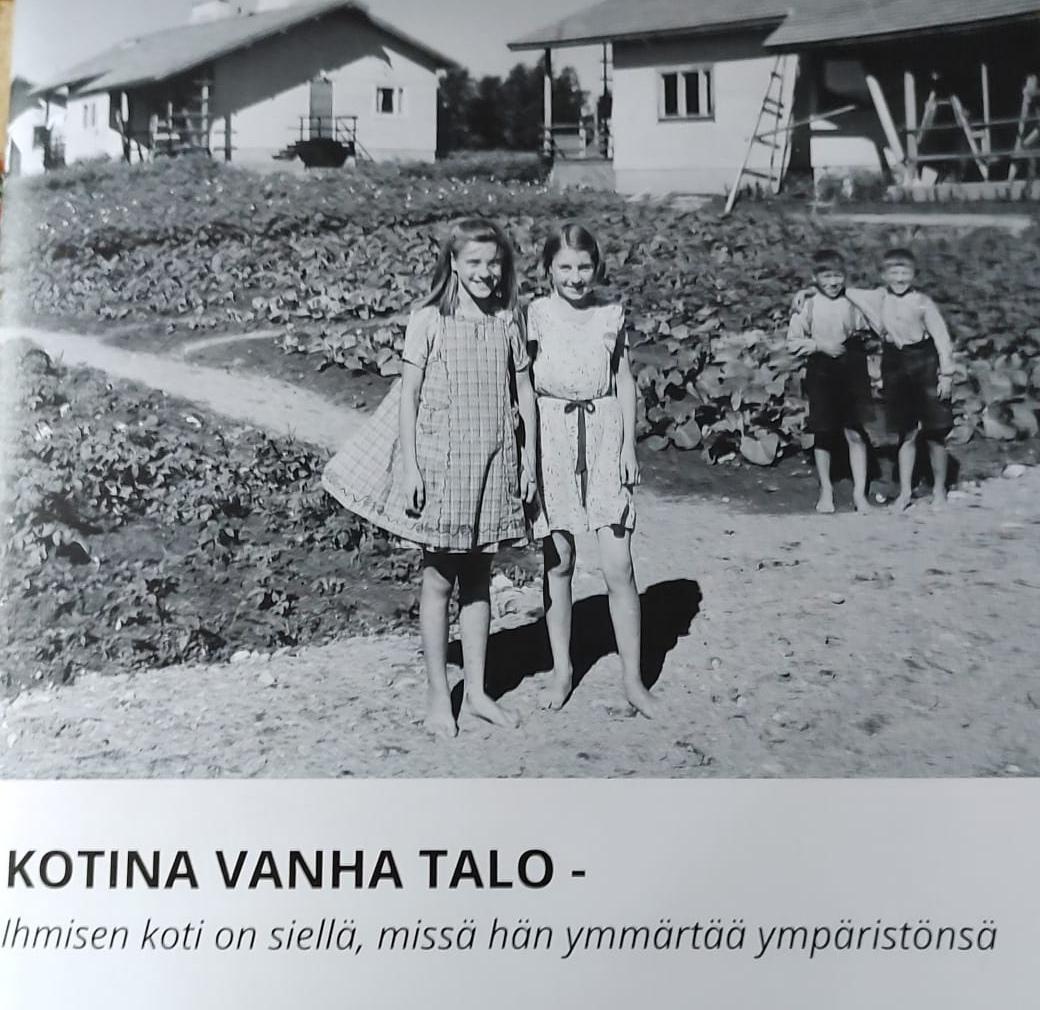 Mustavalkoinen vanha kuva, jossa kaksi tyttöä seisoo puutalon edessä. 