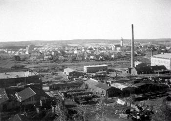 Mustavalkoinen valokuva, jossa näkyy laaja maisema, etualalla tiilisiä teollisuusrakennuksai ja kauempana asutusta. 