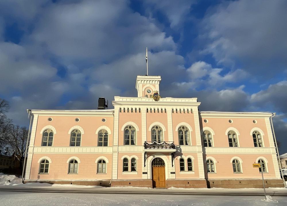 En stor byggnad i rosa med klocktornet och Lovisa fana i toppen. Tvåvåningshus med 18 fönster. Över huvudingången i mitten av byggnaden finns balkong och ovanpå det i takåsen Lovisa vapen.