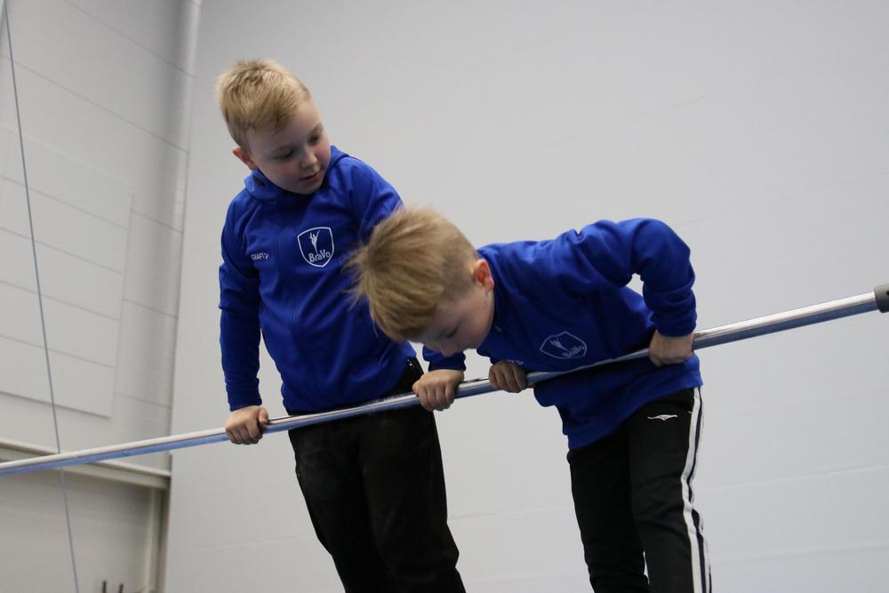Kuvassa on kaksi lasta, jotka ovat pukeutuneet samanlaisiin sinisiin takkeihin, joissa lukee “Bravo”, sekä mustiin housuihin. He ovat sisäliikuntatilassa, jossa on korkea katto ja vaaleat seinäpaneelit. Lapset nojaavat vaakasuoraan metallitankoon ja pitävät siitä kiinni käsillään.
The image shows two children wearing matching blue jackets with the word “Bravo” on them, along with black pants. They are in an indoor sports hall with a high ceiling and light-colored paneled walls. The children are leaning over a horizontal metal bar, gripping it with their hands.