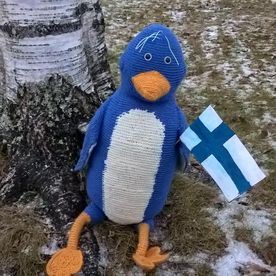 Virkattu sini-valkoinen pääskymaskotti Paltsi alkutalven nurmikolla Suomen lipun kera.