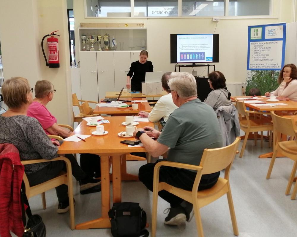 Yhdistyksen salissa on kymmenkunta henkilöä istumassa kahvilla, kuuntelemassa tietoiskua ja keskustelemassa.