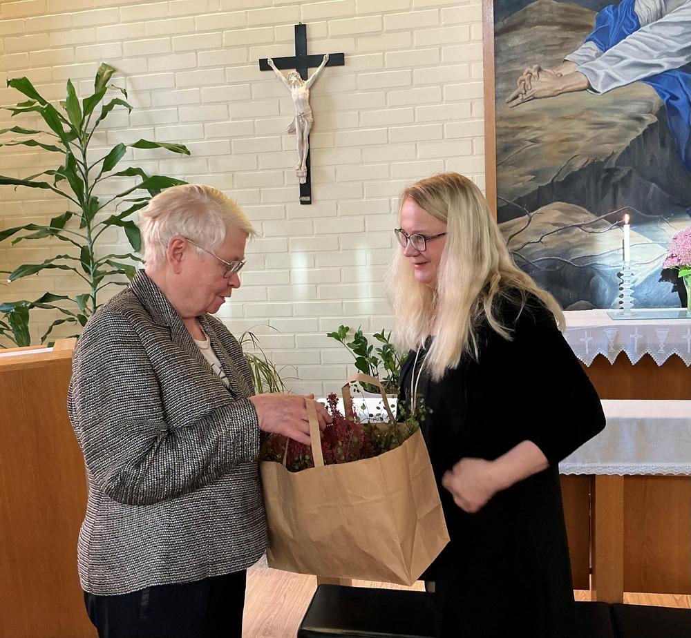 Kaksi iloista vaalea hiuksista naista kohtaavat ja toinen antaa toiselle paperikassin jossa on kukkasia.