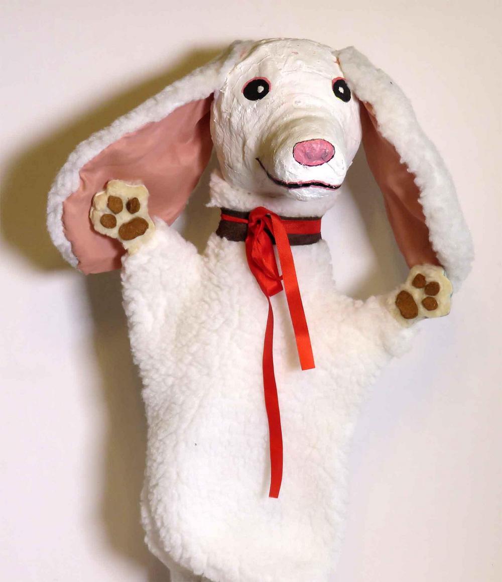 Valkoinen koira-käsinukke. Koiralla on pitkät luppakorvat ja kaulassaan punainen silkkinauha.