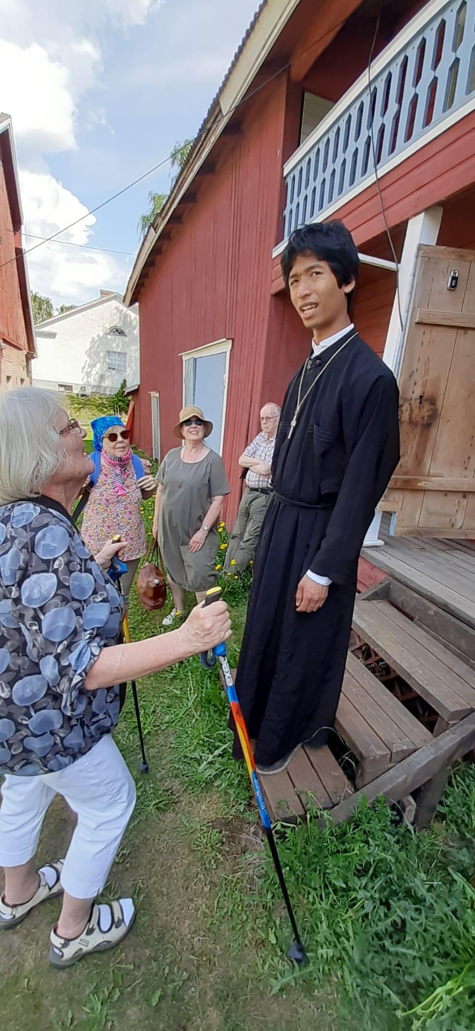 Ihmisiä ulkona kesäpäivänä. Eläkeläisiä ja yksin nuori mies mustassa pitkässä asussa ja risti kaulassaan