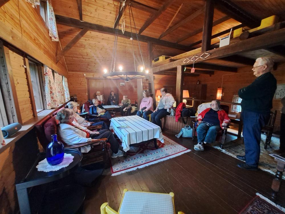 Kolme eläkeläistä kurkistavat tintamareskin, kuvaseinän, reiistä. Maalatussa tintamareskissa henkilöillä on kansallispuvut yllään.