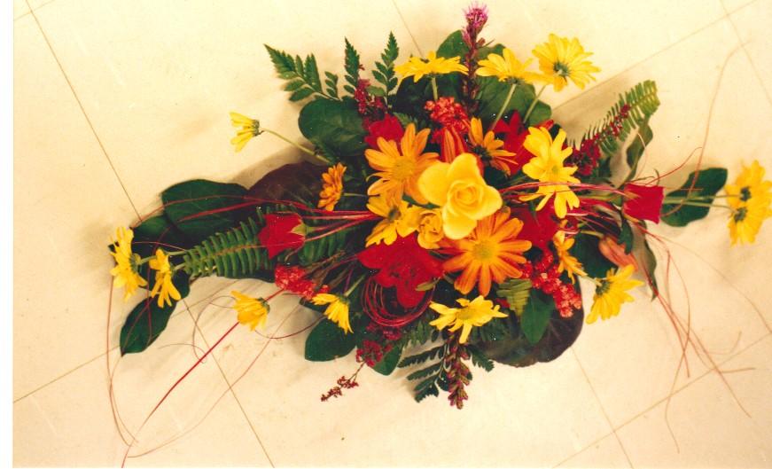 Kukka-asetelma jossa on keltaisia, punaisia ja oransseja kukkia, sekä myös vihreitä lehtiä