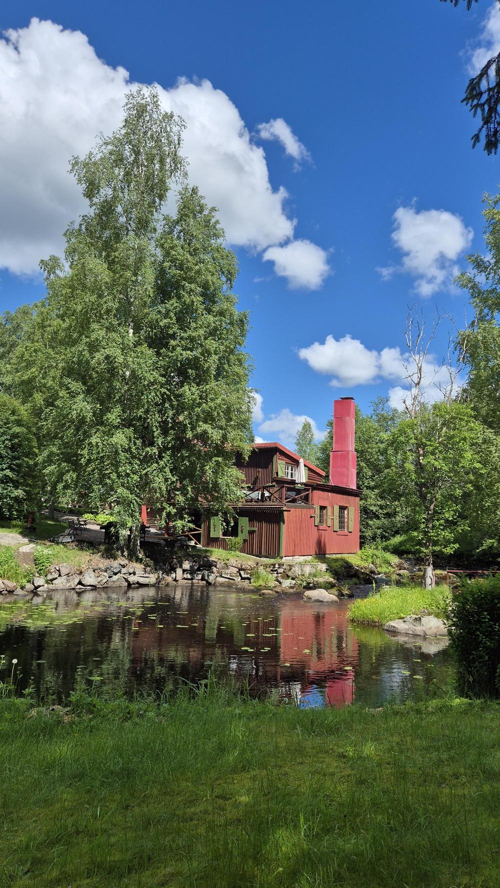 Aurinkoinen ja kesäinen maisema. Joen äärellä on punainen puurakennus, mylly. Rakennuksen vieressä on suuri koivu, taustalla sininen taivas.