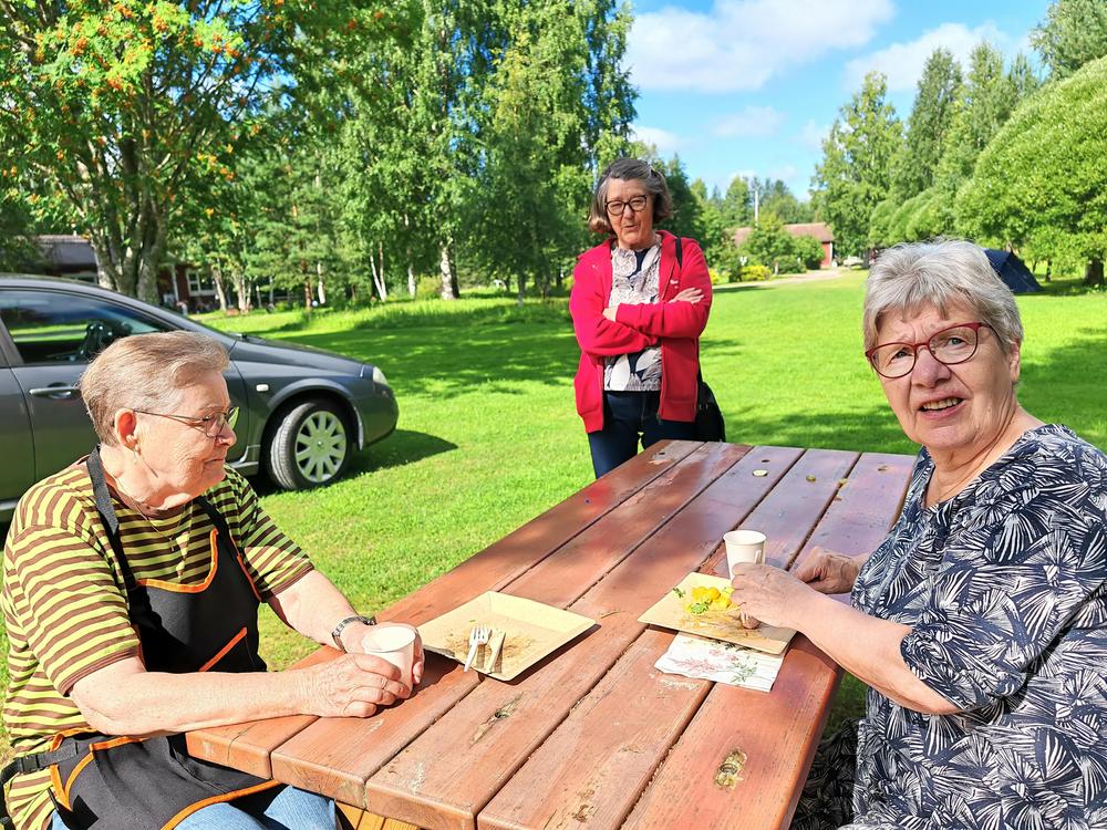 Kolme naista ulkona, kaksi istuu pöydän ääressä ja  yksi seisoo pöydän vieressä