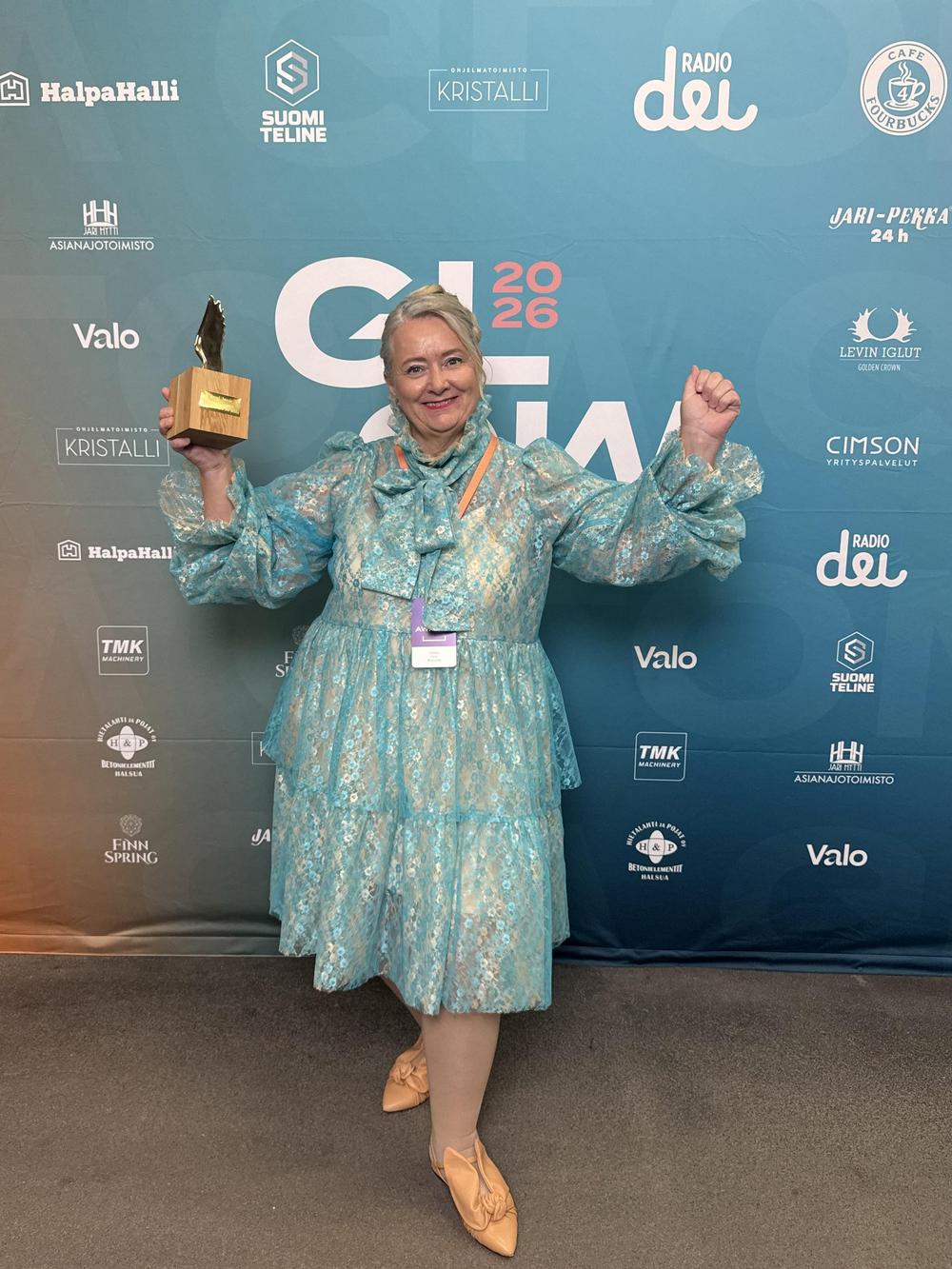 Juhlapukuisella Kirsi Rostamolla on palkinto kädessään ja hän hymyilee kameralle. Hänen takanaan mediaseinällä on Glow Festivaalin sponsoreiden logoja.