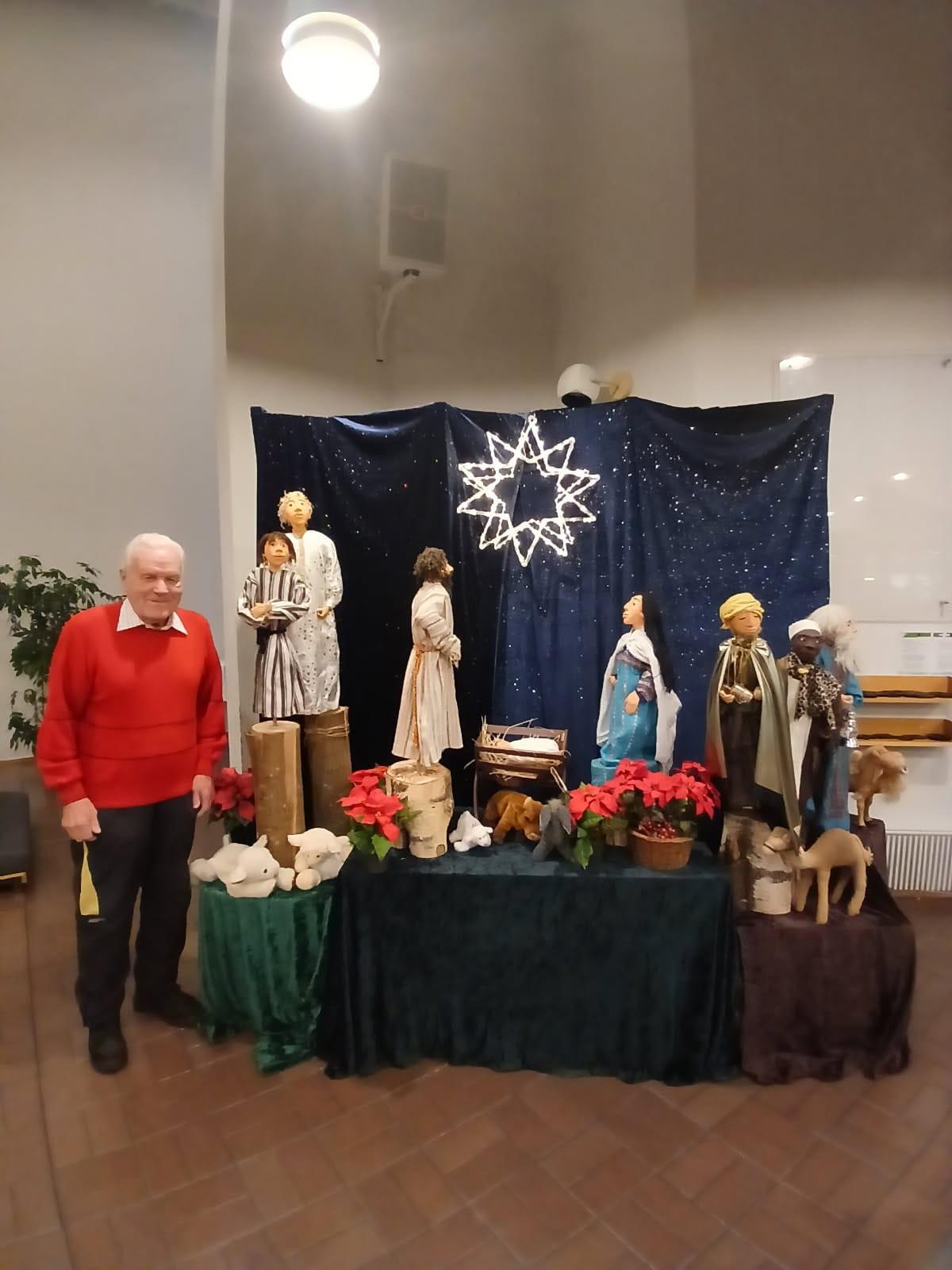 Hymyilevä eläkeläismies punaisessa paidassa seisoo seimikuvaelman vieressä seurakuntatalon sisällä.