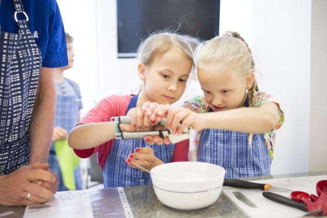 Kaksi vaaleaa marttaessuihin sonnustautunutta tyttöä puuhaa keittiössä aikuisen seuratessa vierestä.
