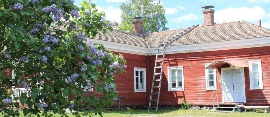 Kuvassa punamullan värinen museorakennus ja sen edessä kukkivat syreenit 