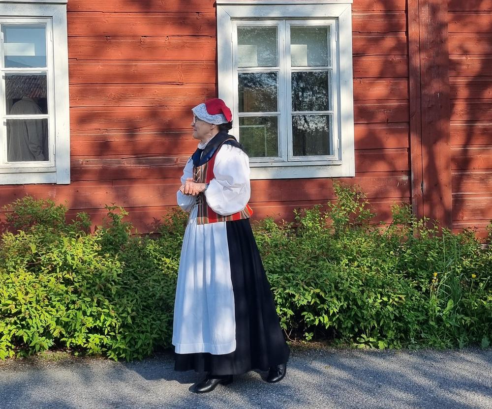 Nainen päällään Maskun seudun kansallispuvun värikäs liivi, pitkä hame ja esiliina punamultaisen Maskun museorakennuksen edustalla