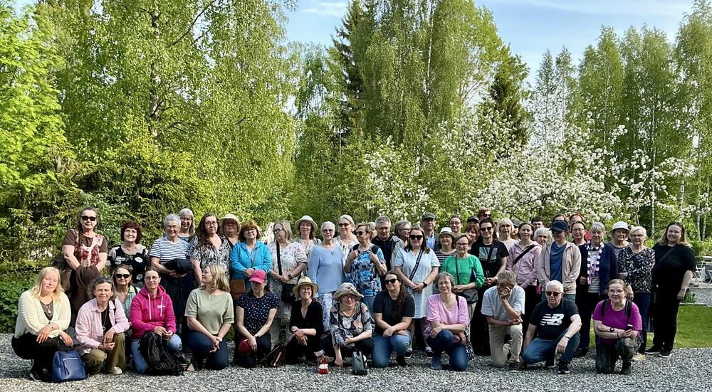 Ryhmäkuva kesäisessä maisemassa. Kuvan etualalla on noin 50 henkilön seurue. Henkilöt katsovat kuvaajaa iloisin ilmein. Kuvan taustalla on koivuja ja kukkiva omenapuu.