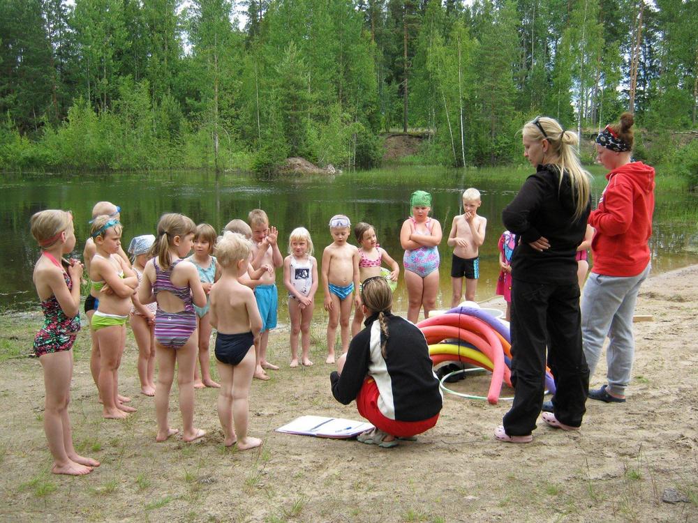 Taka-alalla lampi, etualalla joukko lapsia ja kolme aikuista hiekkarannalla. 