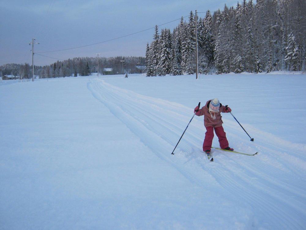 Luminen peltomaisema, jota halkoo latu. Ladulla lapsi hiihtämässä. 