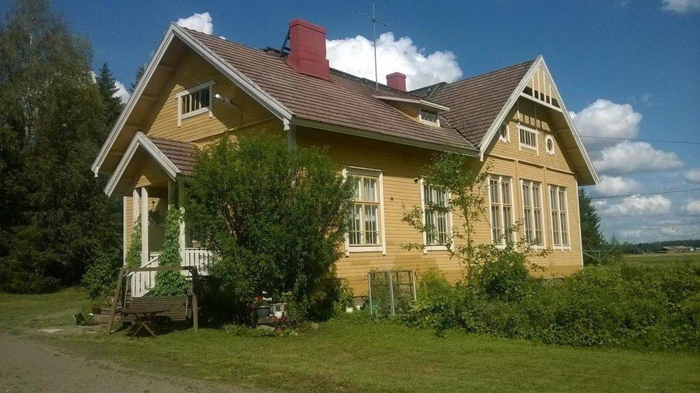 Vaaleankeltainen puutalo, ruskea katto, valkoiset karmit, kesäinen, vihreä ympäristö. 