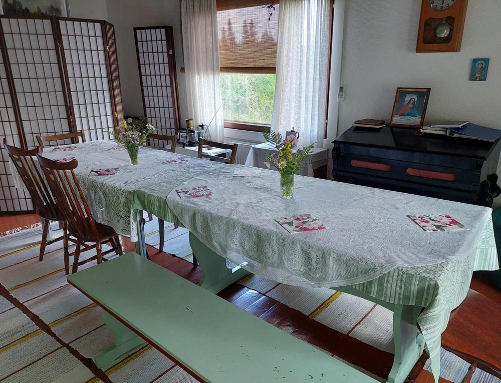 Tuvassa ruokapöytä, jolla on valkeat pitsiliinat. Lattialla on räsymatot.