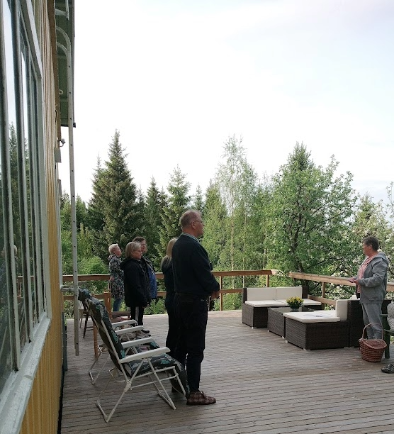 Hiljaisuuden retriitin osanottajat seisovat talon terassilla ja kuuntelevat puhujaa.