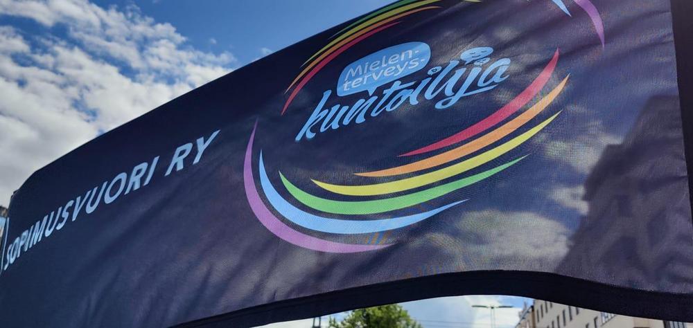 Sopimusvuori ry:n tumma banderolli, jossa lukee Sopimusvuori ry ja Mielenterveyskuntoilija. Lisäksi sateenkaaren väreissä oleva pyöreä kuvio.