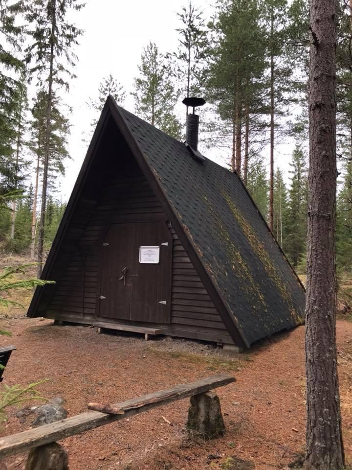 Tummanpuhuva, A-mallinen kotarakennus harvennetun puuston keskellä metsässä. Kodan ovet on suljettu ja ympäristö siisti.